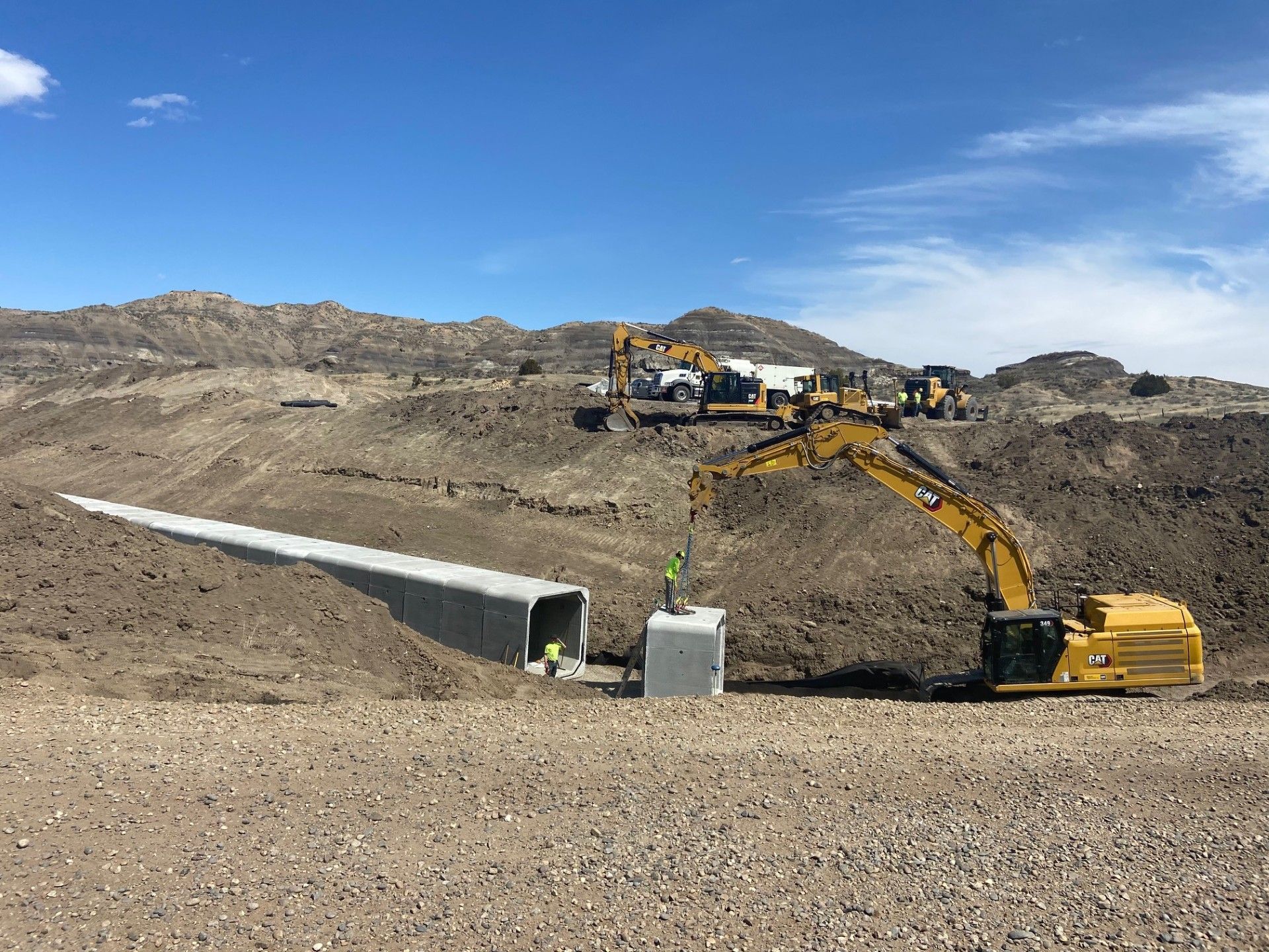 Construction site with excavator placing a concrete box culvert in a dirt landscape, mountains in the background.