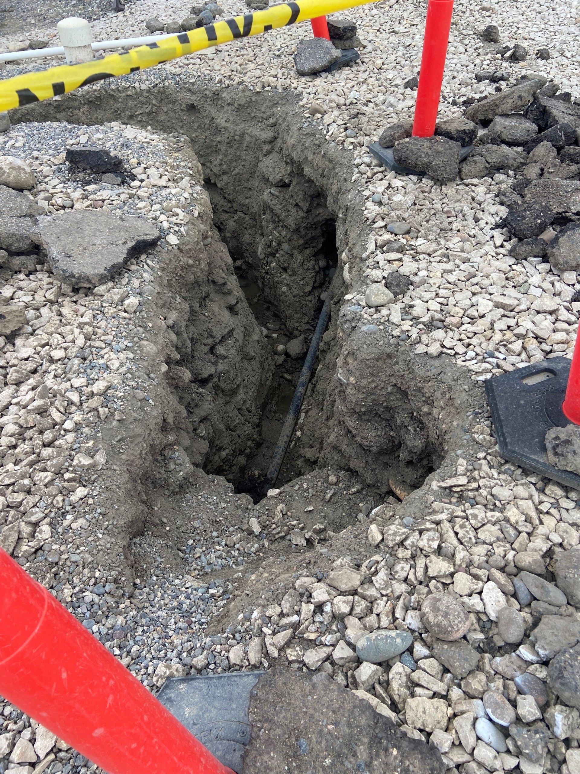 A narrow trench dug in gravel and asphalt, with caution tape and red poles marking the area.