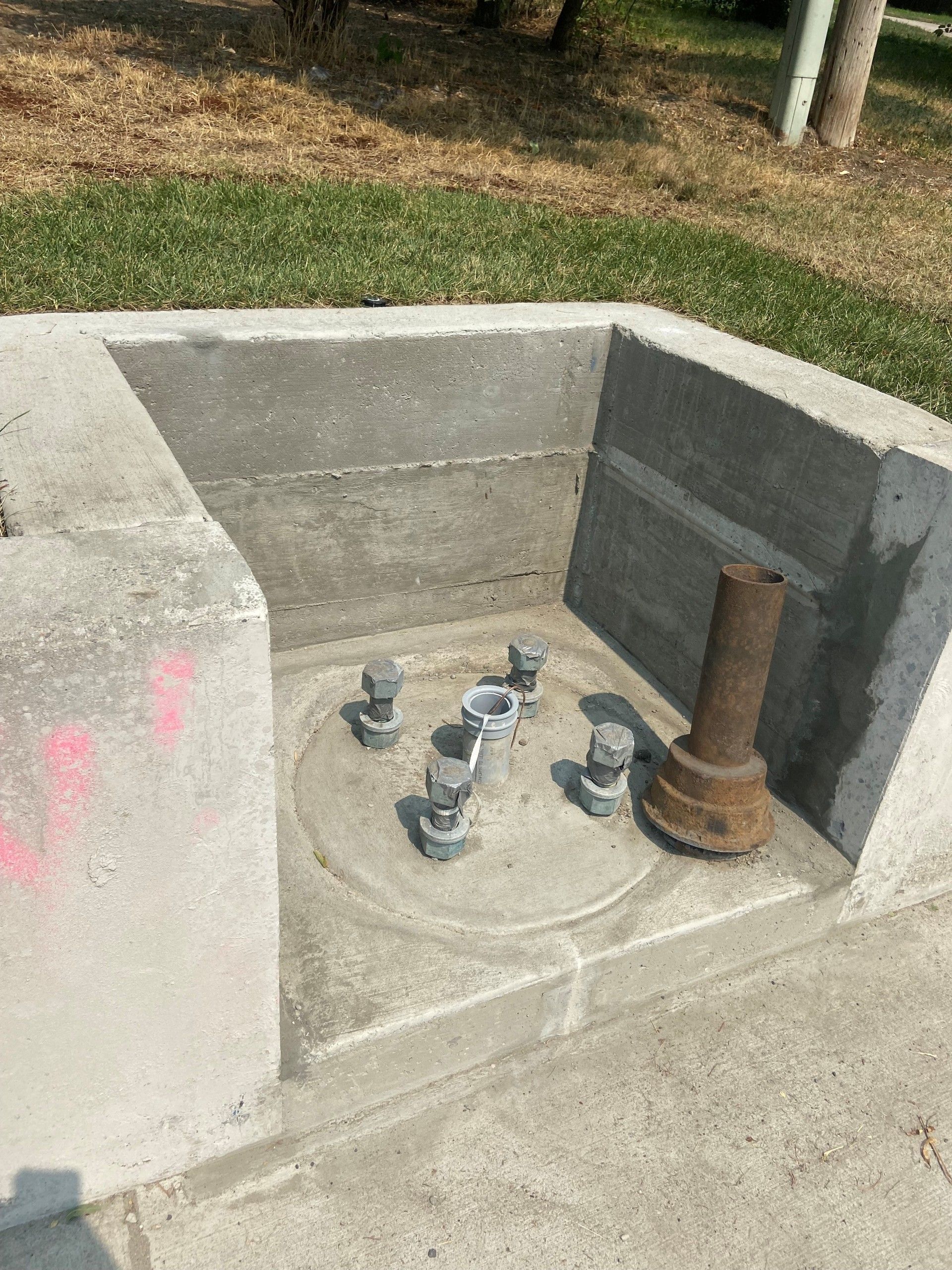 Concrete foundation with bolts and a metal post, likely for a light pole. Outdoors, sunny day.
