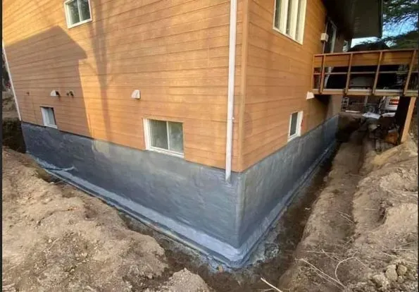 Fondations de la maison recouvertes d'un mastic foncé, tranchées apparentes à la base. Bardage en bois au-dessus.