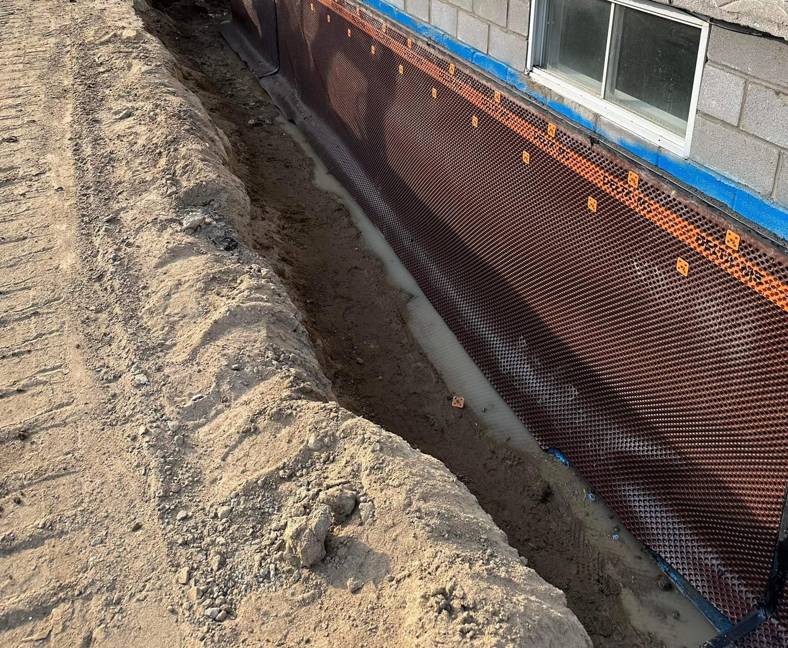 Tranchée creusée à côté des fondations d'un bâtiment, laissant apparaître une membrane de drainage contre le mur et de la terre meuble.