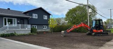 Une petite pelleteuse orange sur un terrain en terre battue, devant une maison.