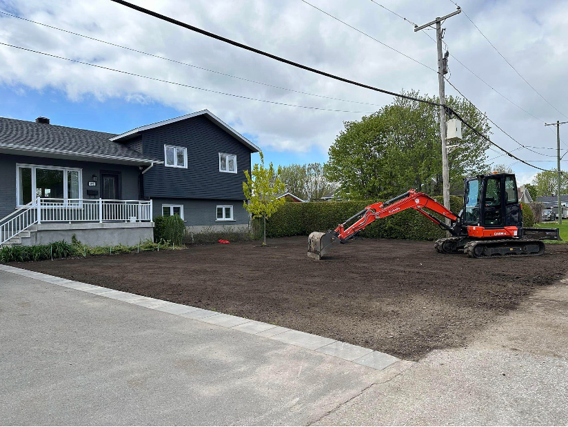 Une excavatrice rouge travaille sur un terrain en terre devant une maison.