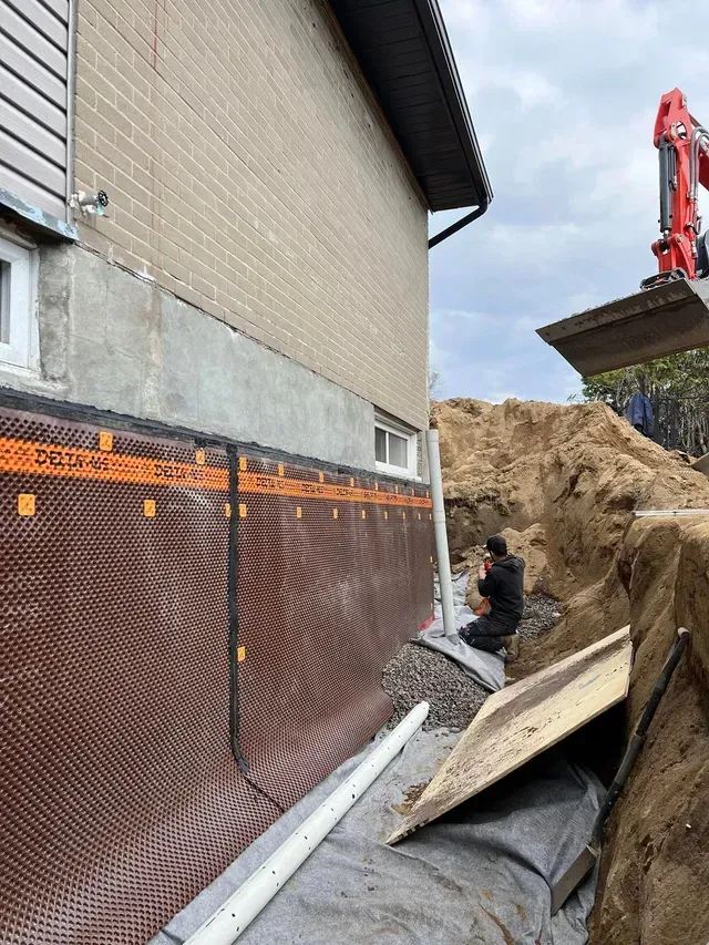 Travaux de fondations extérieures de la maison avec pose d'une membrane de drainage. Une excavatrice et un ouvrier sont présents.
