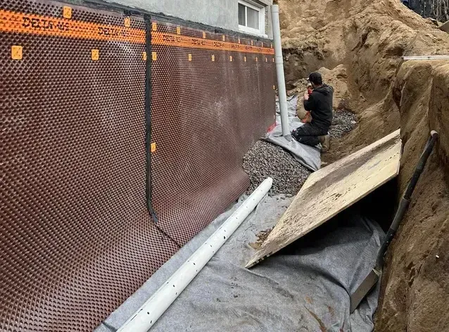 Un homme installe une membrane de drainage brune sur un mur de fondation en béton, à côté d'une tranchée.