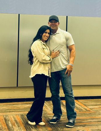 A woman in black pants and a tan shirt stands with a man in jeans and a gray shirt, posing together.