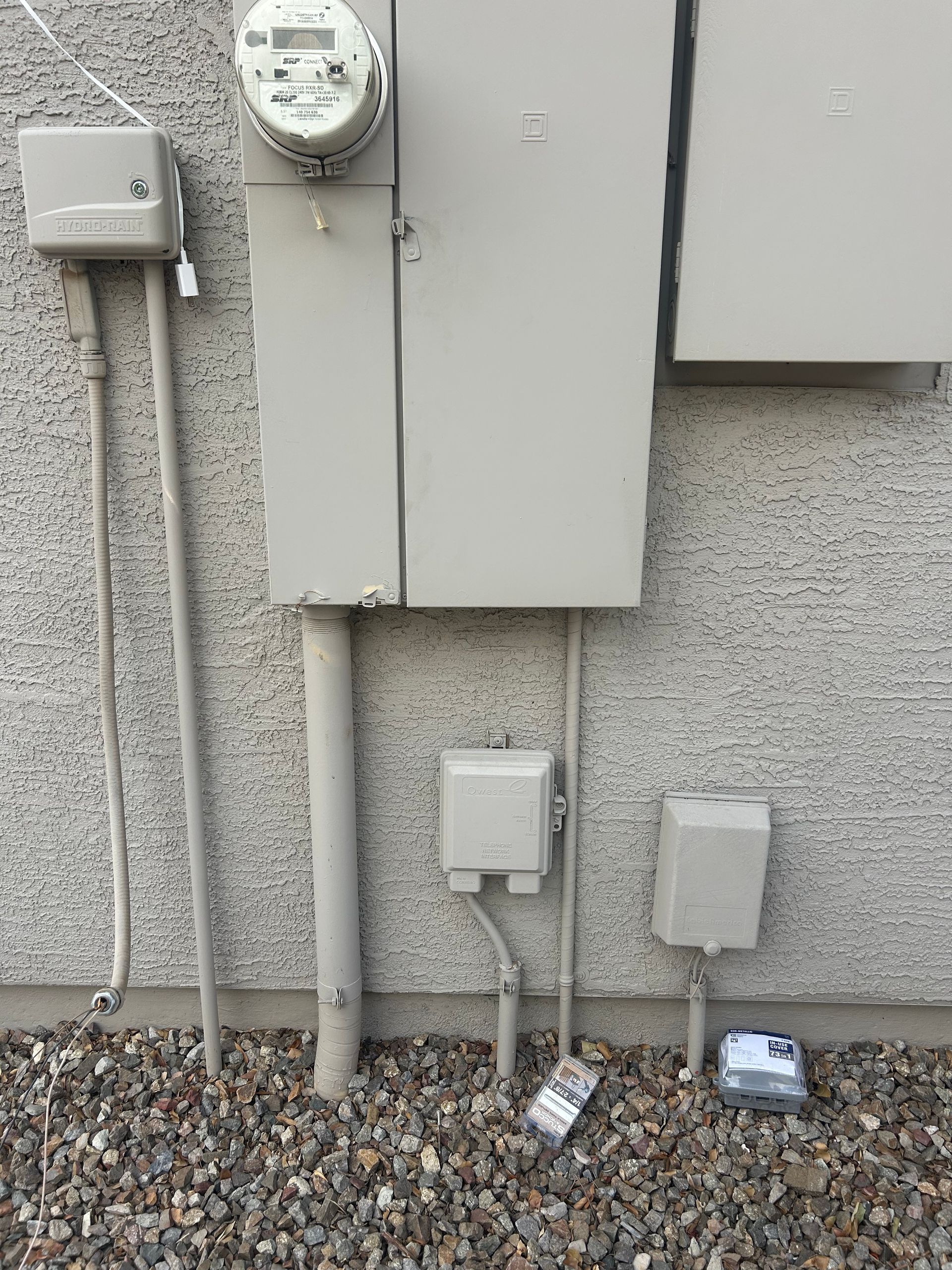 Electrical equipment on a stucco wall: meter, boxes, conduits, and small service boxes on gravel.