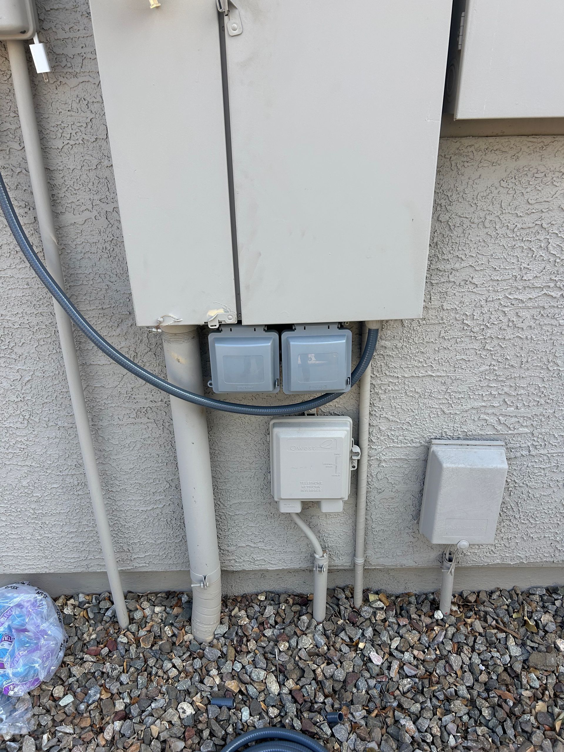 Exterior electrical boxes on a stucco wall. Gray conduit and wiring. Gravel ground.