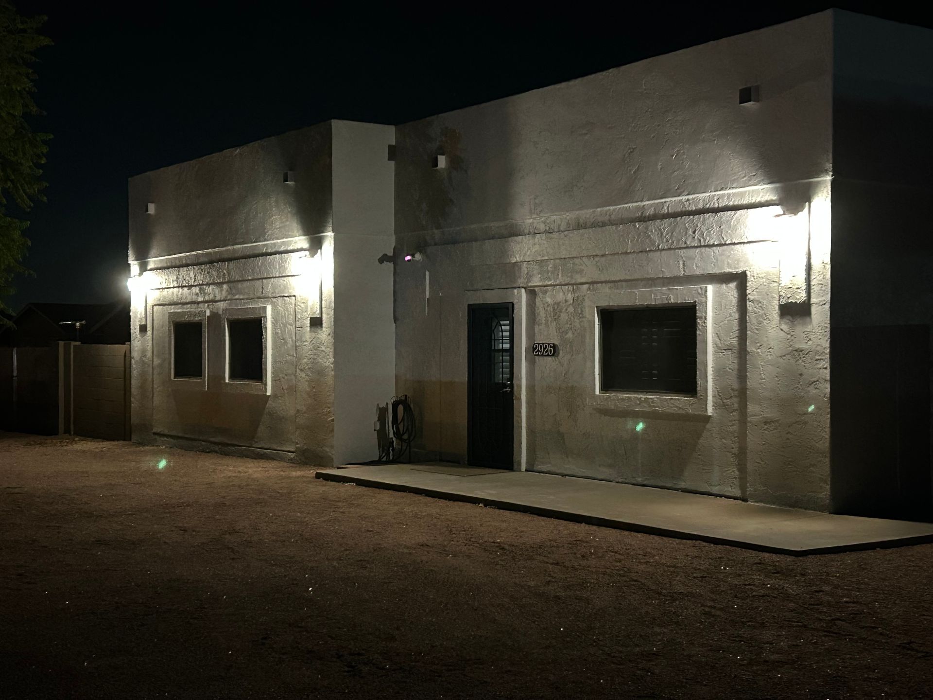 Building exterior at night, lit by wall sconces. Gravel ground. Dark windows and door.