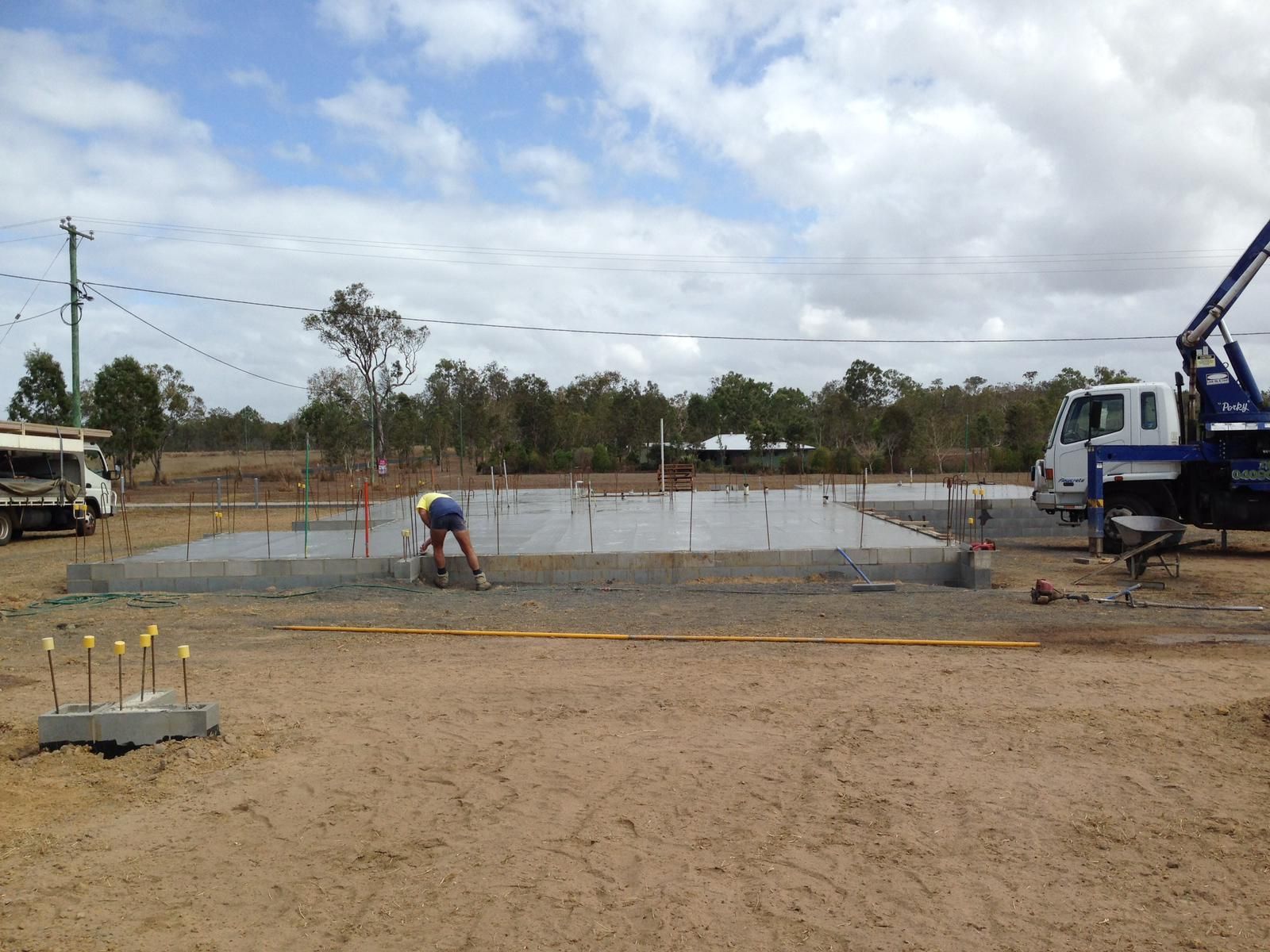 Concrete foundation being poured at a construction site; worker leveling surface. Trees in background, cloudy sky.