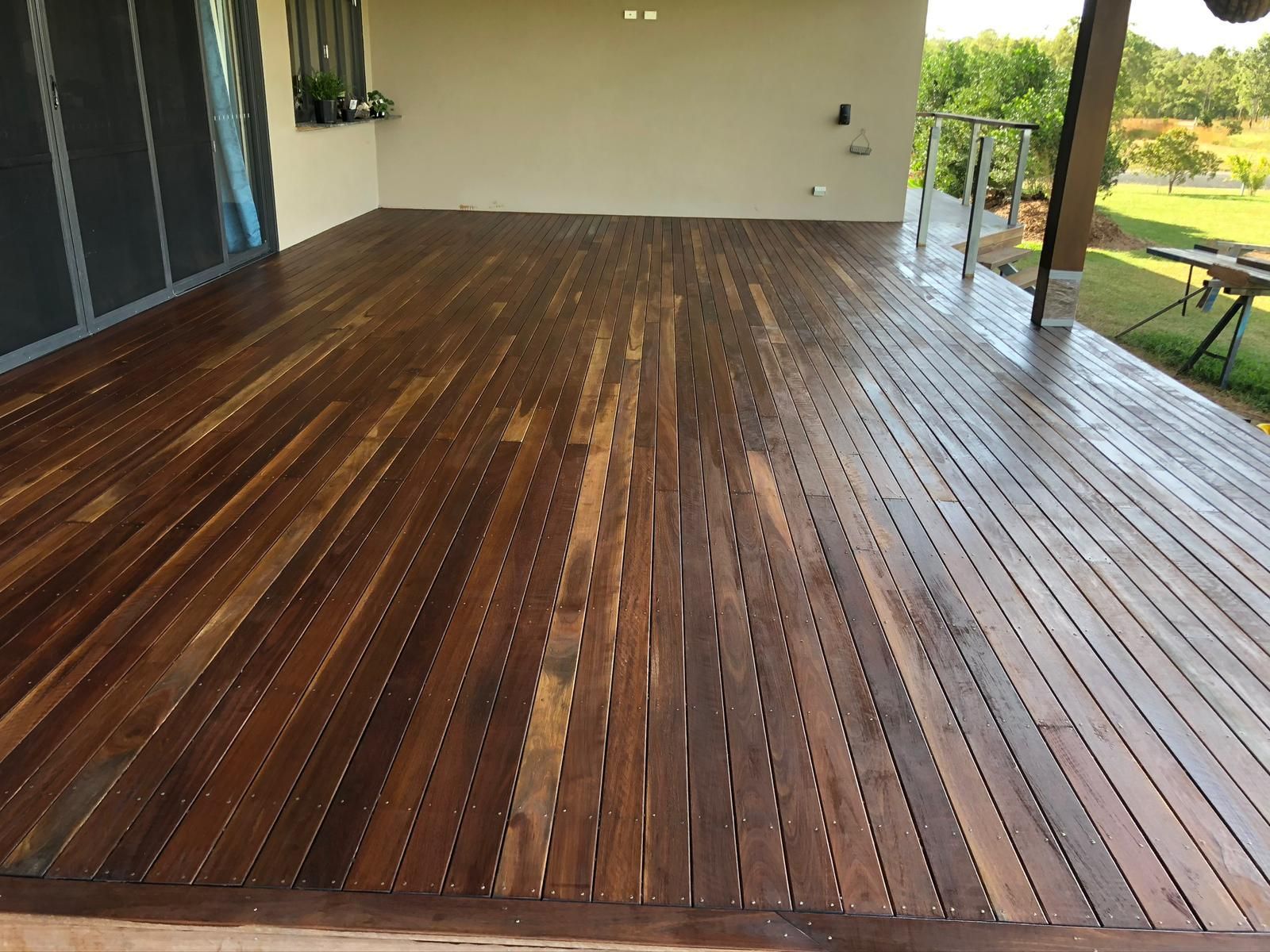 Wooden deck with wet, dark-stained boards under a covered patio, with a glimpse of a grassy landscape.