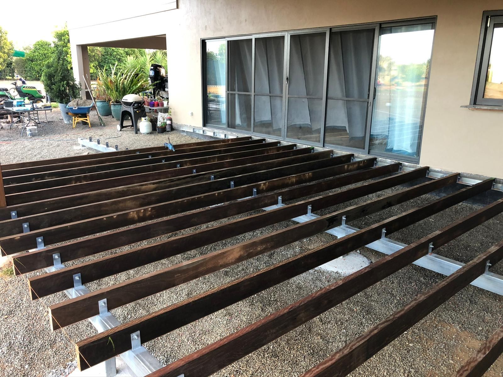 Wooden deck under construction, with horizontal beams supported by metal brackets on gravel.