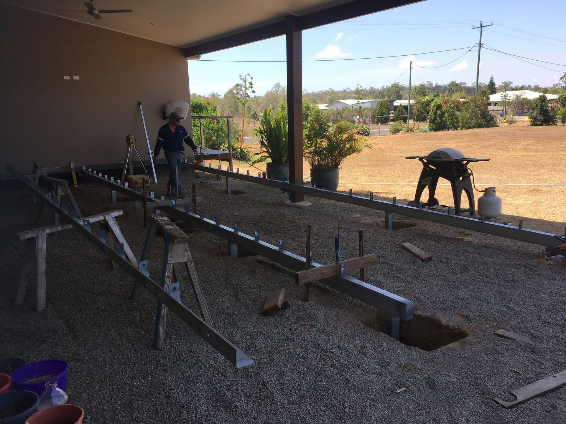 Construction of a deck on a gravel surface. Two people working, metal beams and tools present. Brown surroundings.