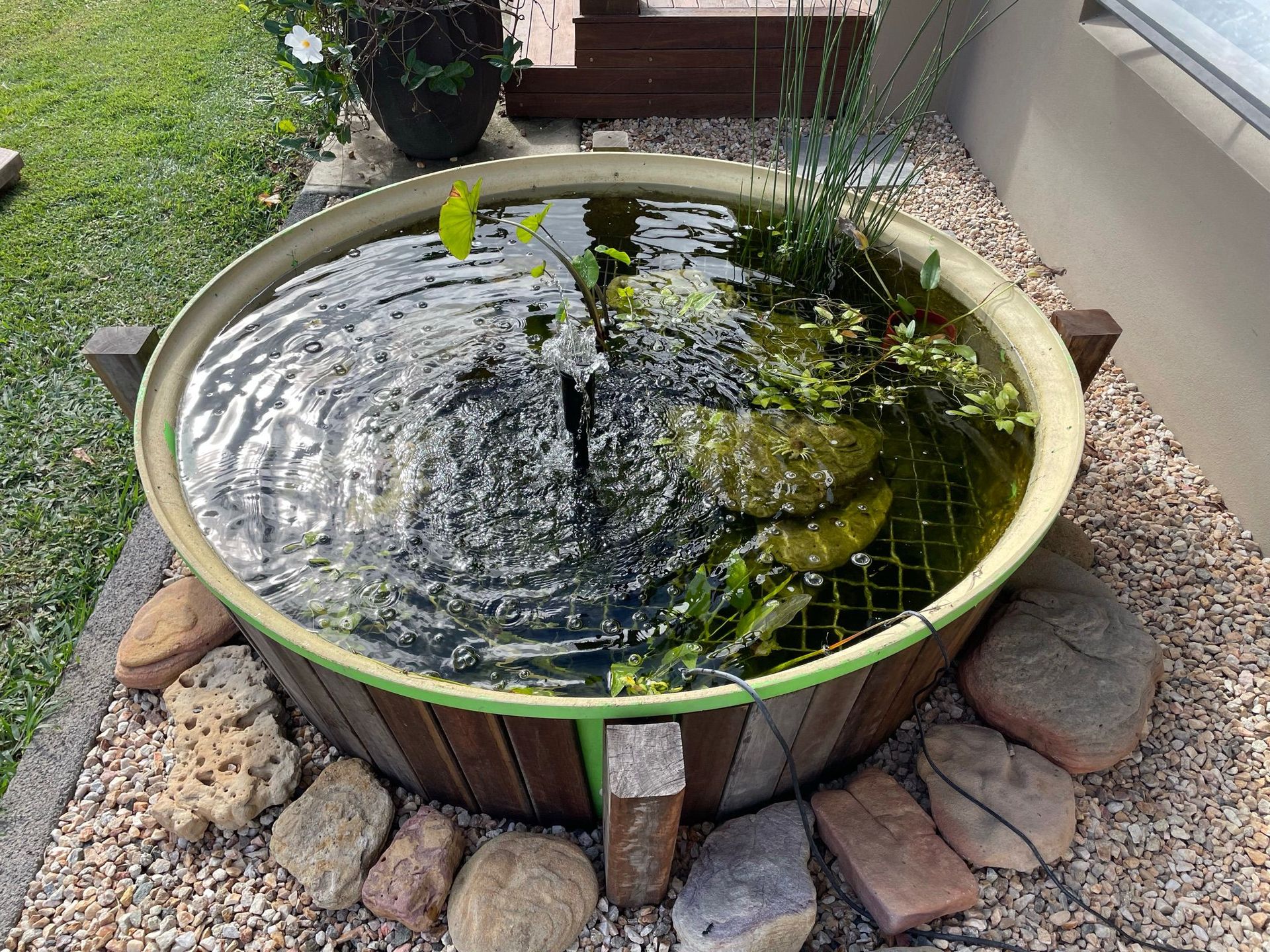 Circular outdoor pond with rippling water, plants, rocks, and gravel.