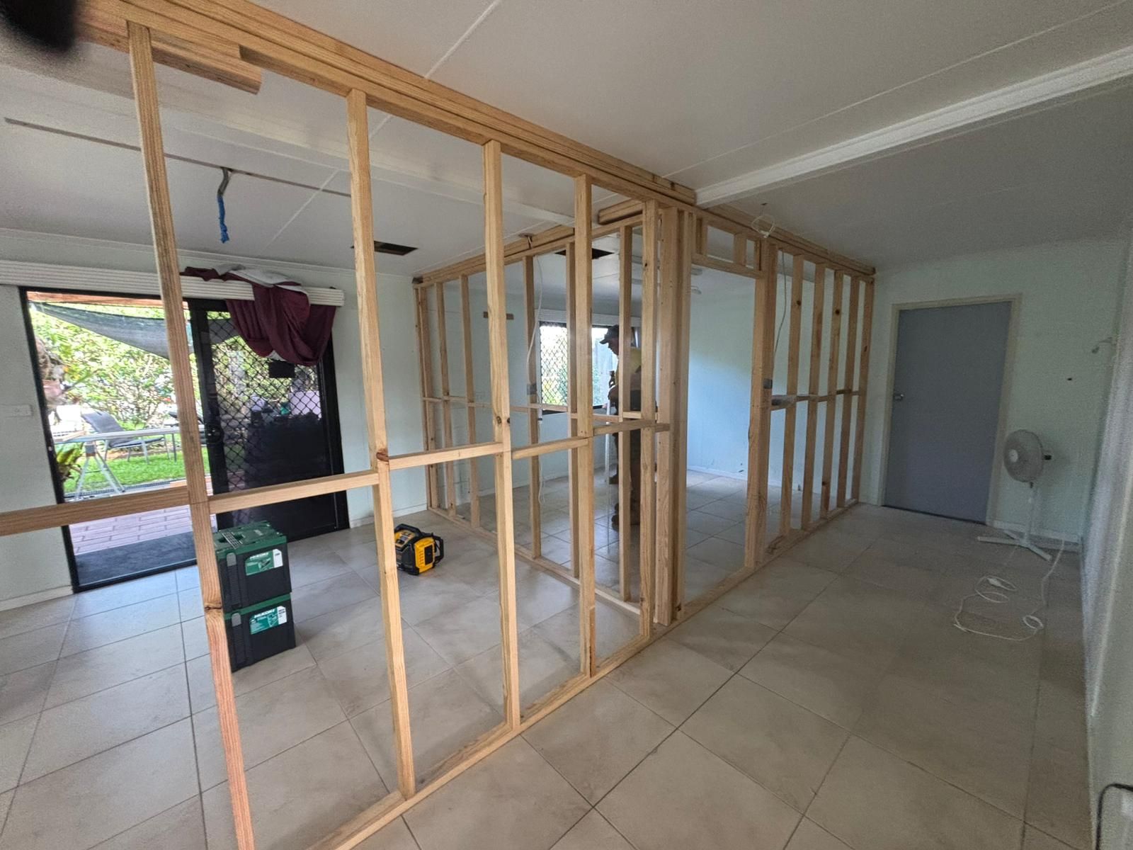 Interior renovation: Wooden wall framing underway in a room with a doorway to a backyard, concrete tiled floors.