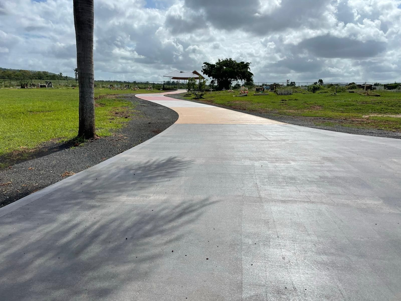 A concrete road curves towards a structure, flanked by green fields under a cloudy sky.