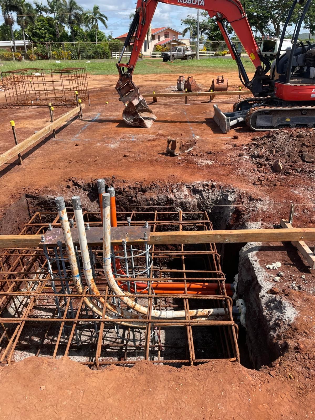 Construction site: Excavator digging near rebar cage with conduits, red soil, and wood framing.
