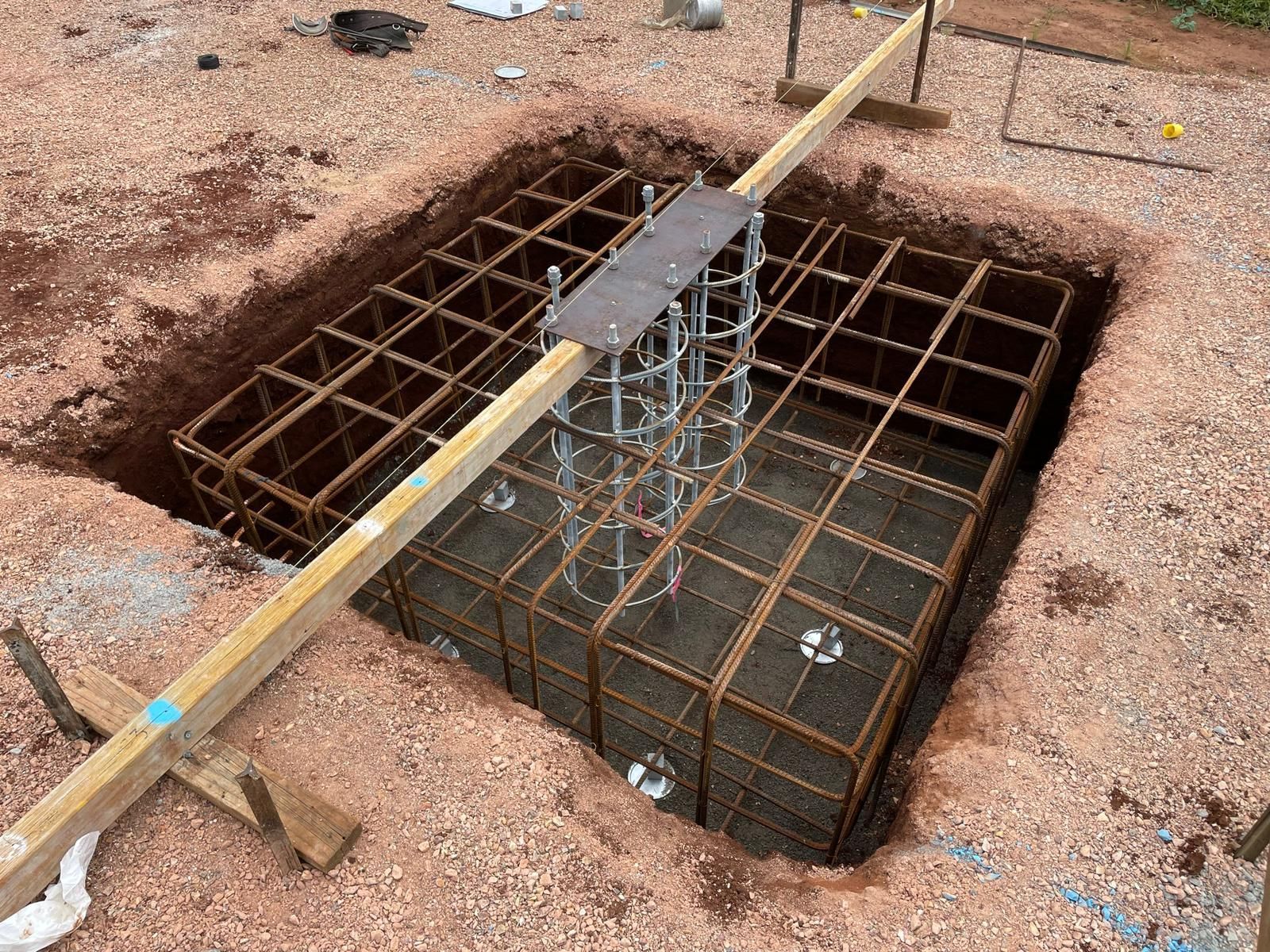 A rectangular foundation pit with steel reinforcement, ready for concrete pour. A wooden beam spans the pit.
