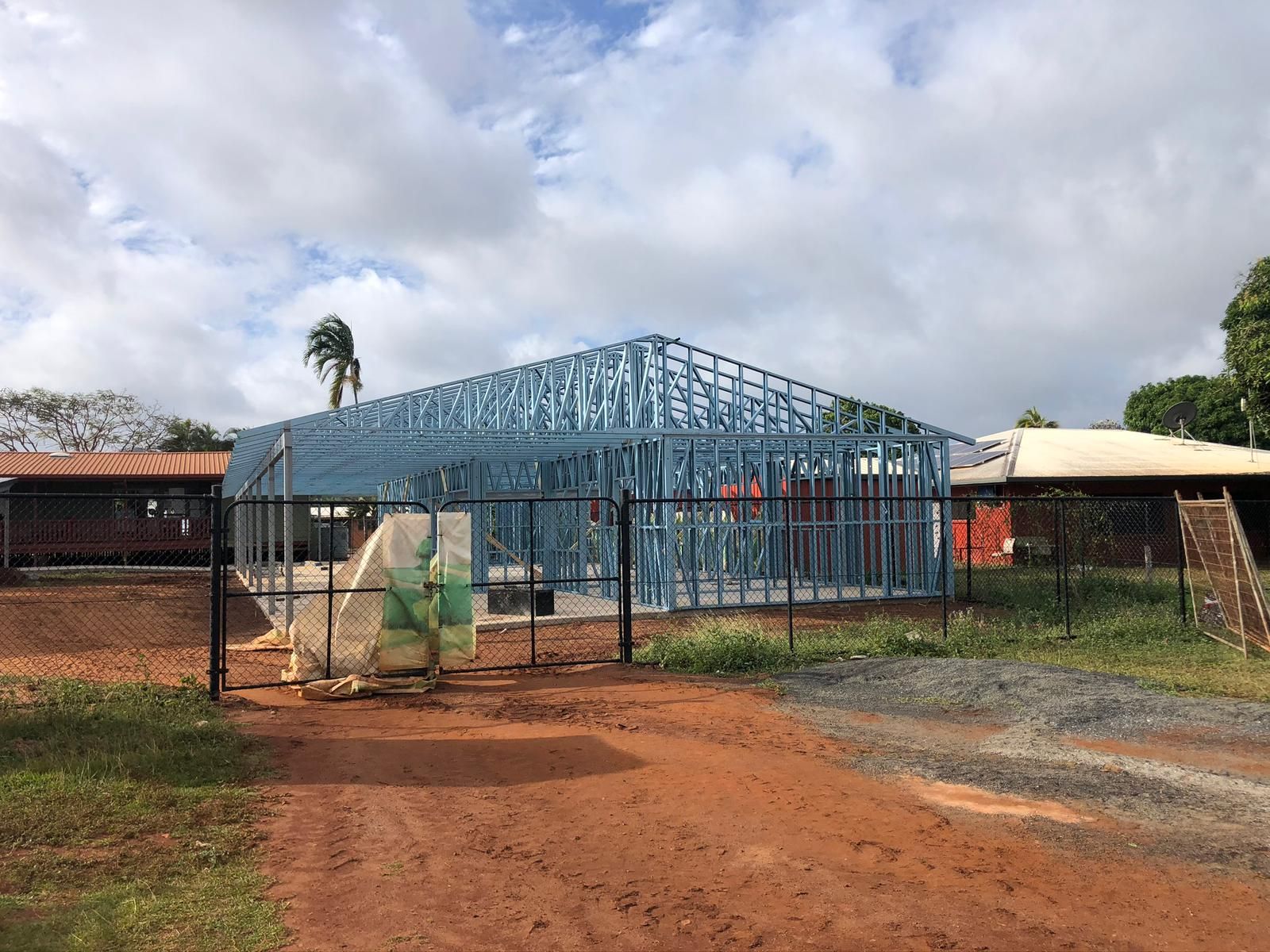 Steel frame of a building under construction, set on a concrete foundation, behind a chain-link fence on a dirt lot.
