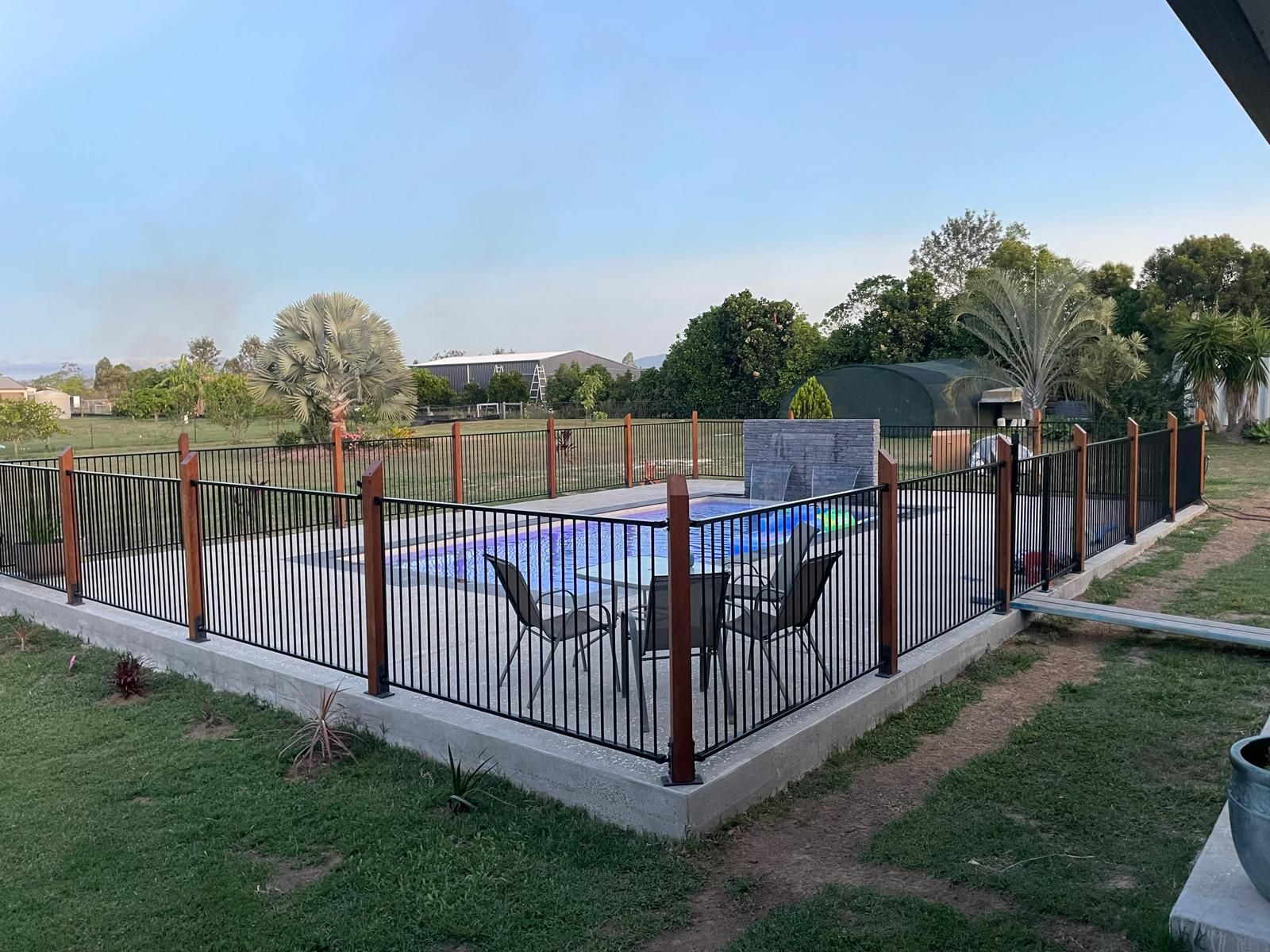 Pool area with black and wood fence on a grassy lawn. Blue tiled pool with chairs inside.