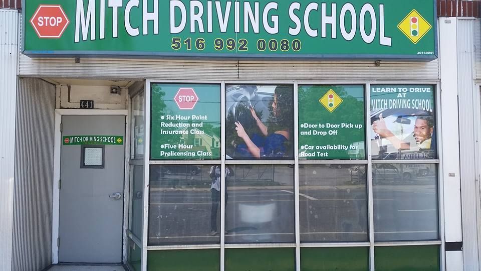 The storefront for Mitch Driving School, featuring a green sign with phone number 516-992-0080 and windows with ads.