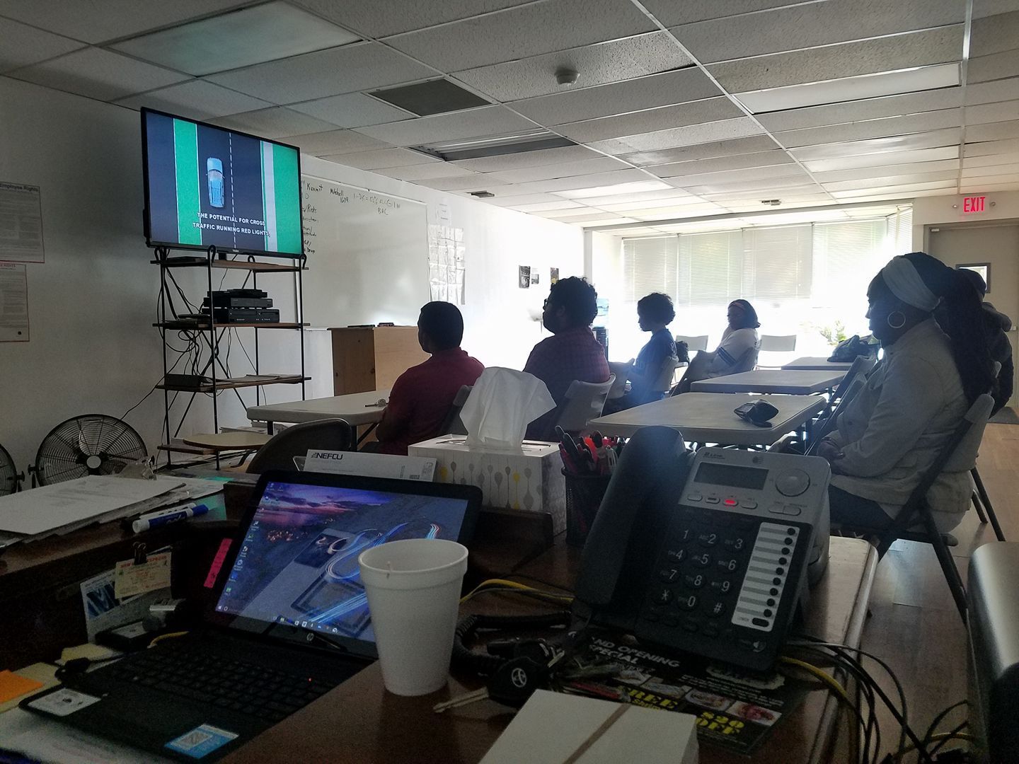 A group of people sitting at tables in a room, facing a screen showing a presentation.