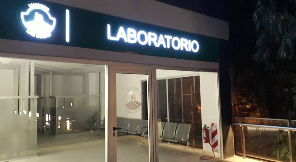 Letrero de entrada al laboratorio iluminado por la noche. Letrero verde y blanco; paredes de cristal y sillas interiores.