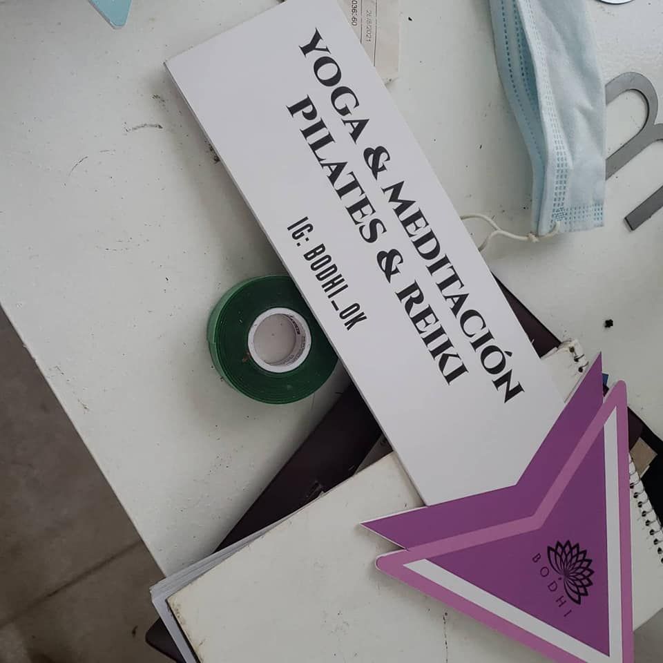 Letrero blanco: Yoga, Pilates, Meditación, Reiki. Flecha morada, rollo de cinta verde y mascarilla sobre una mesa blanca.