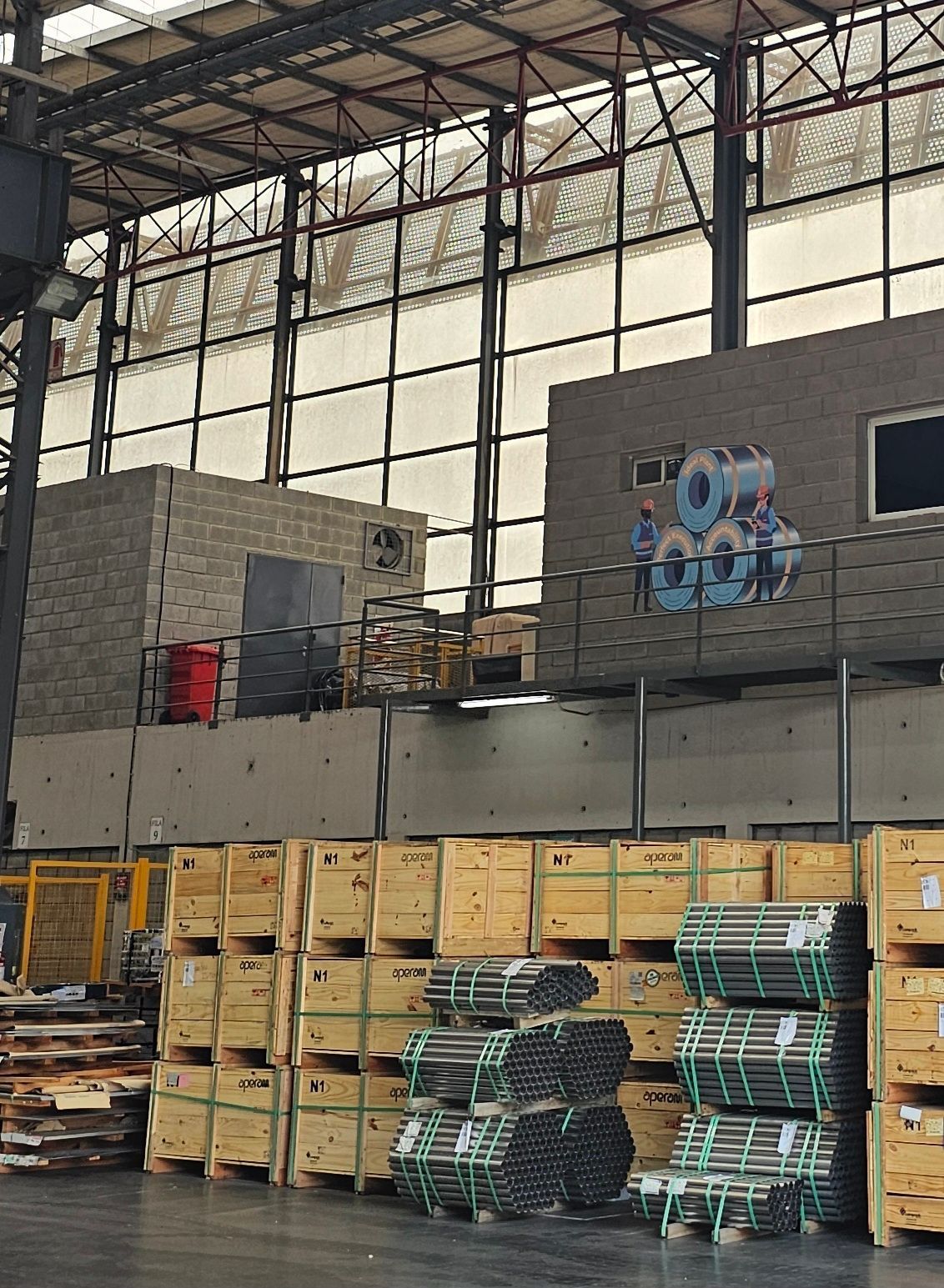 Interior de almacén con cajas de madera apiladas y bobinas de metal; dos estructuras de hormigón al fondo.