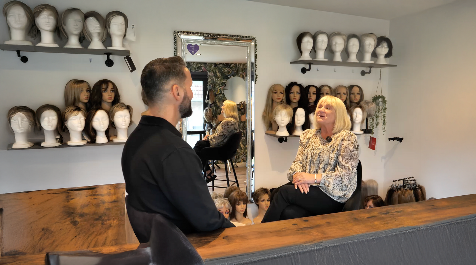 Man facing a woman in a wig shop. Wigs are displayed on shelves. A mirror reflects the shop interior.