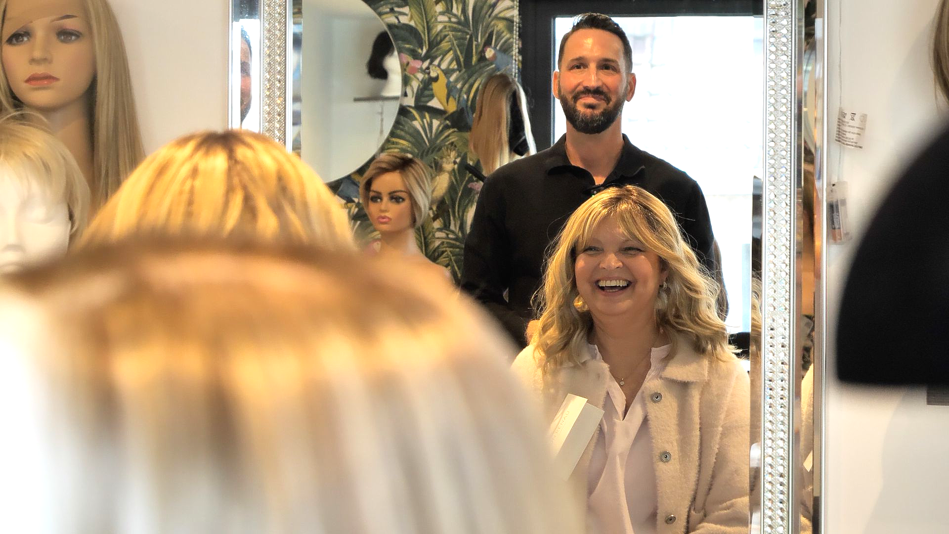 Woman smiles in mirror, reflected in foreground. A man stands behind her. Salon setting with wigs.