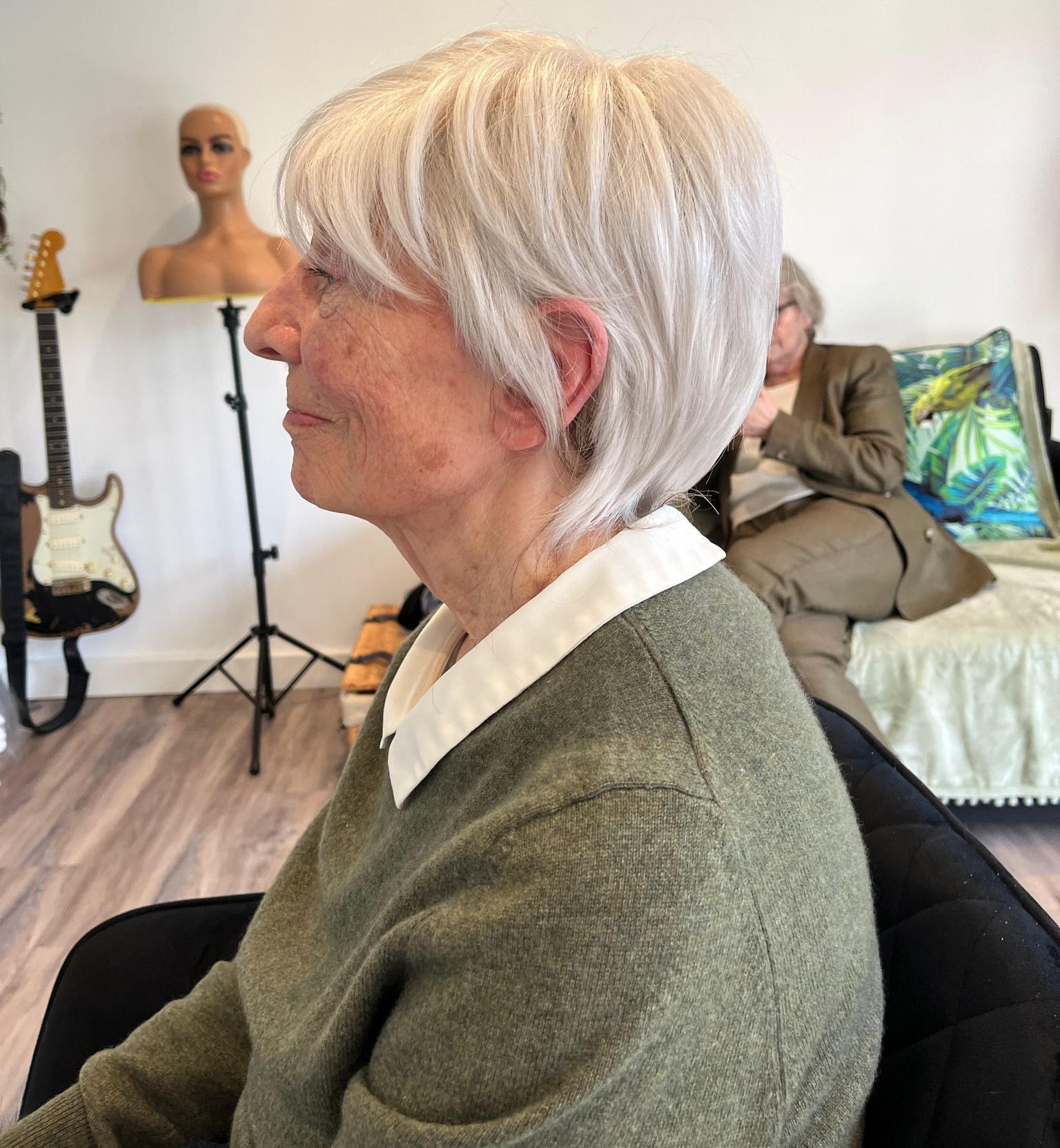 Woman with short, light hair sits in a chair. She wears a green sweater and white collared shirt.