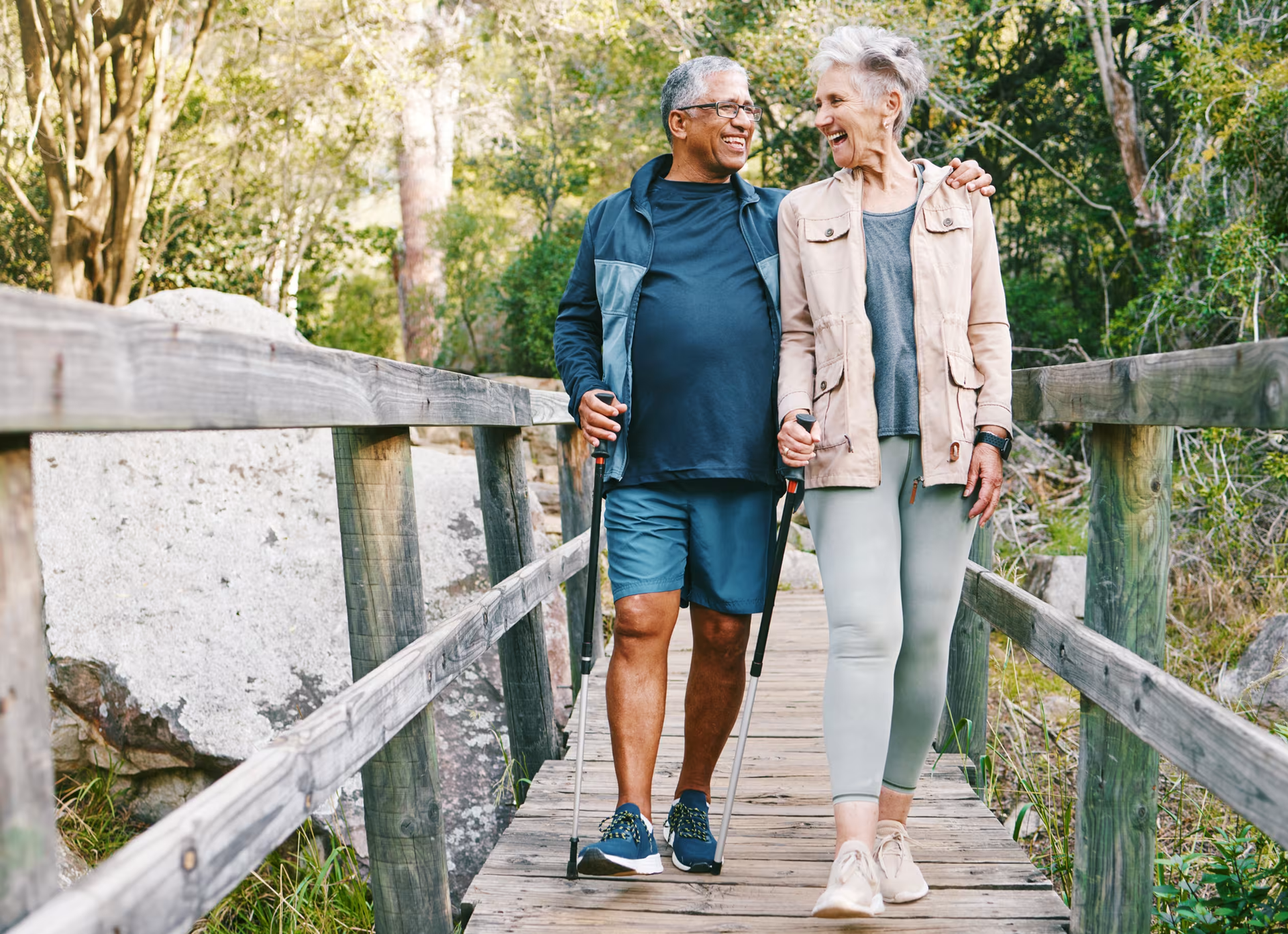 An elderly couple is walking across a wooden bridge in the woods.