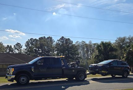 A dark tow truck hauling a black SUV on a sunny street. Trees and houses are in the background.
