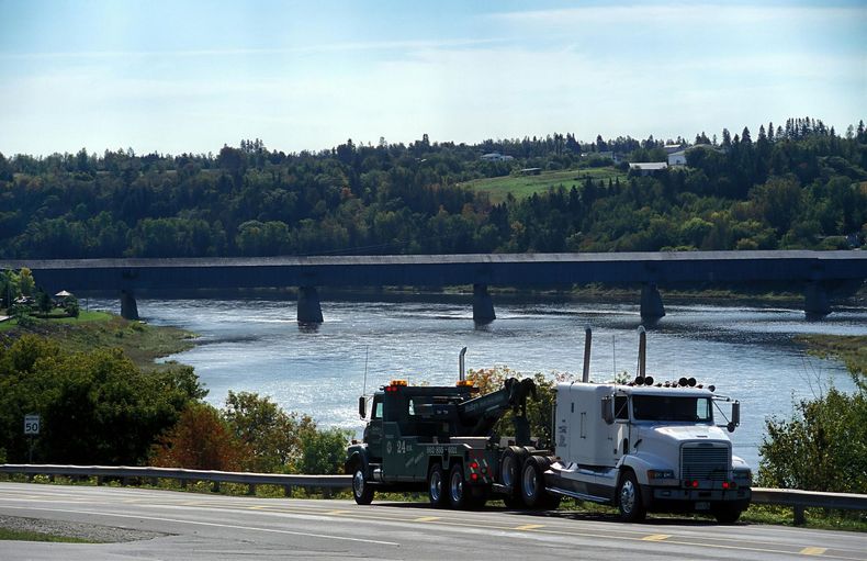 Tow truck pulling a vehicle near a bridge over a river. Green trees and blue sky background.