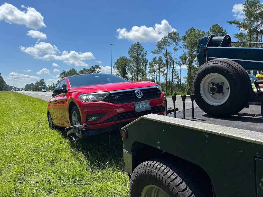 Red car being towed by a tow truck on the side of a highway, in a grassy area, under a blue sky.