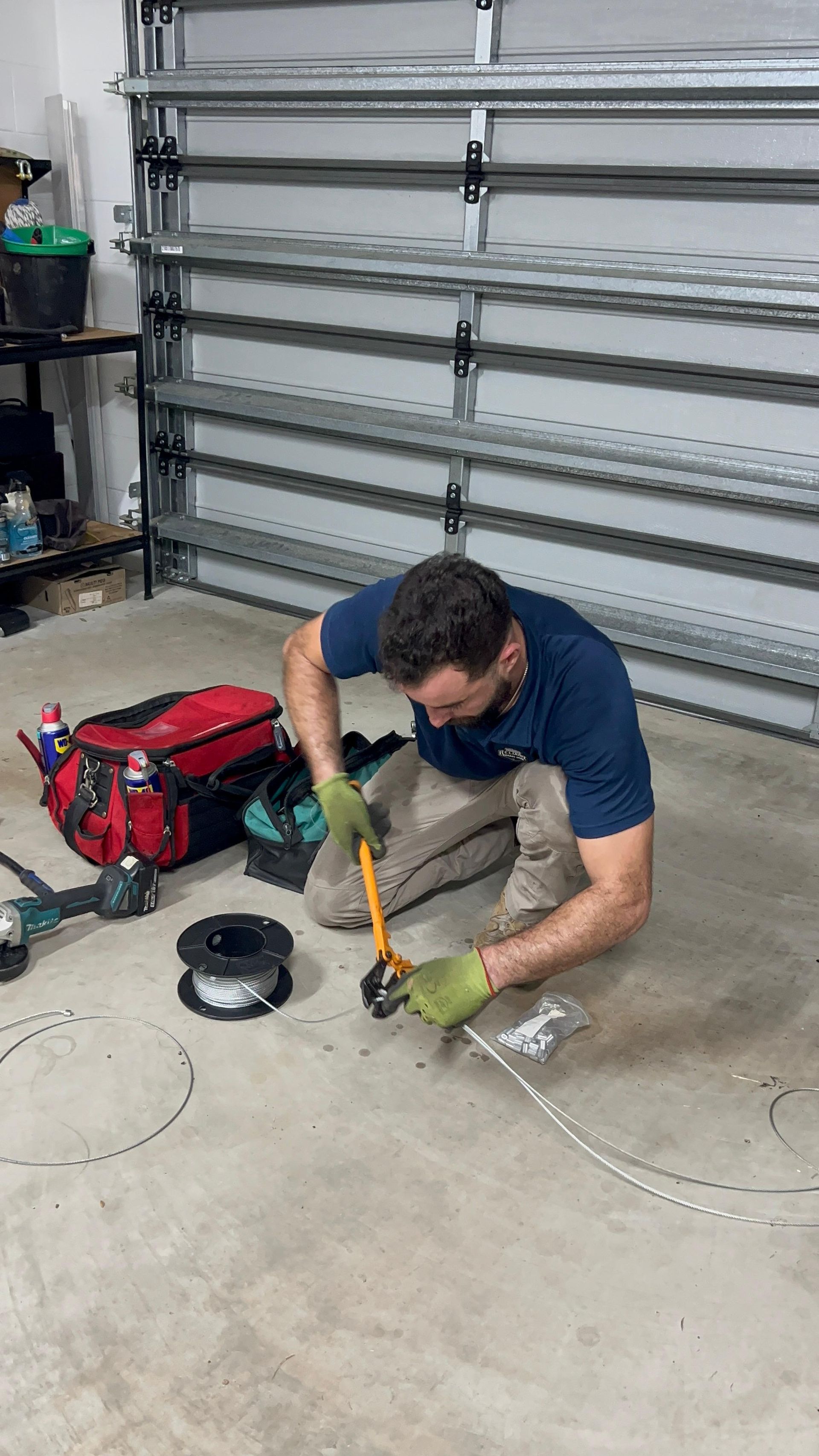 Man kneeling on a garage floor, using a hammer on a metal tool beside a red tool bag and coils.
