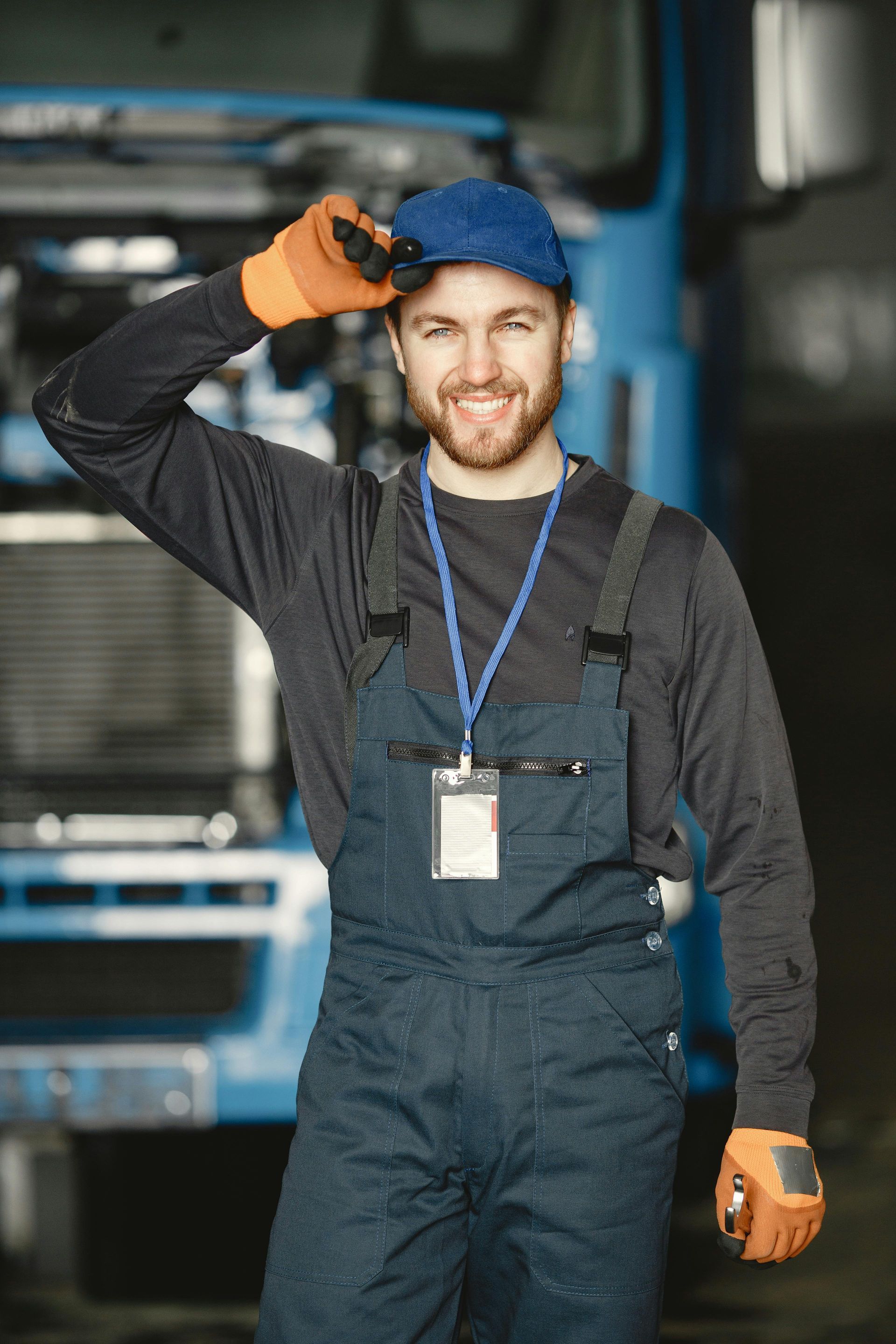 Mechanic in blue overalls and cap smiling, touching cap, orange gloves, in front of a blue truck.