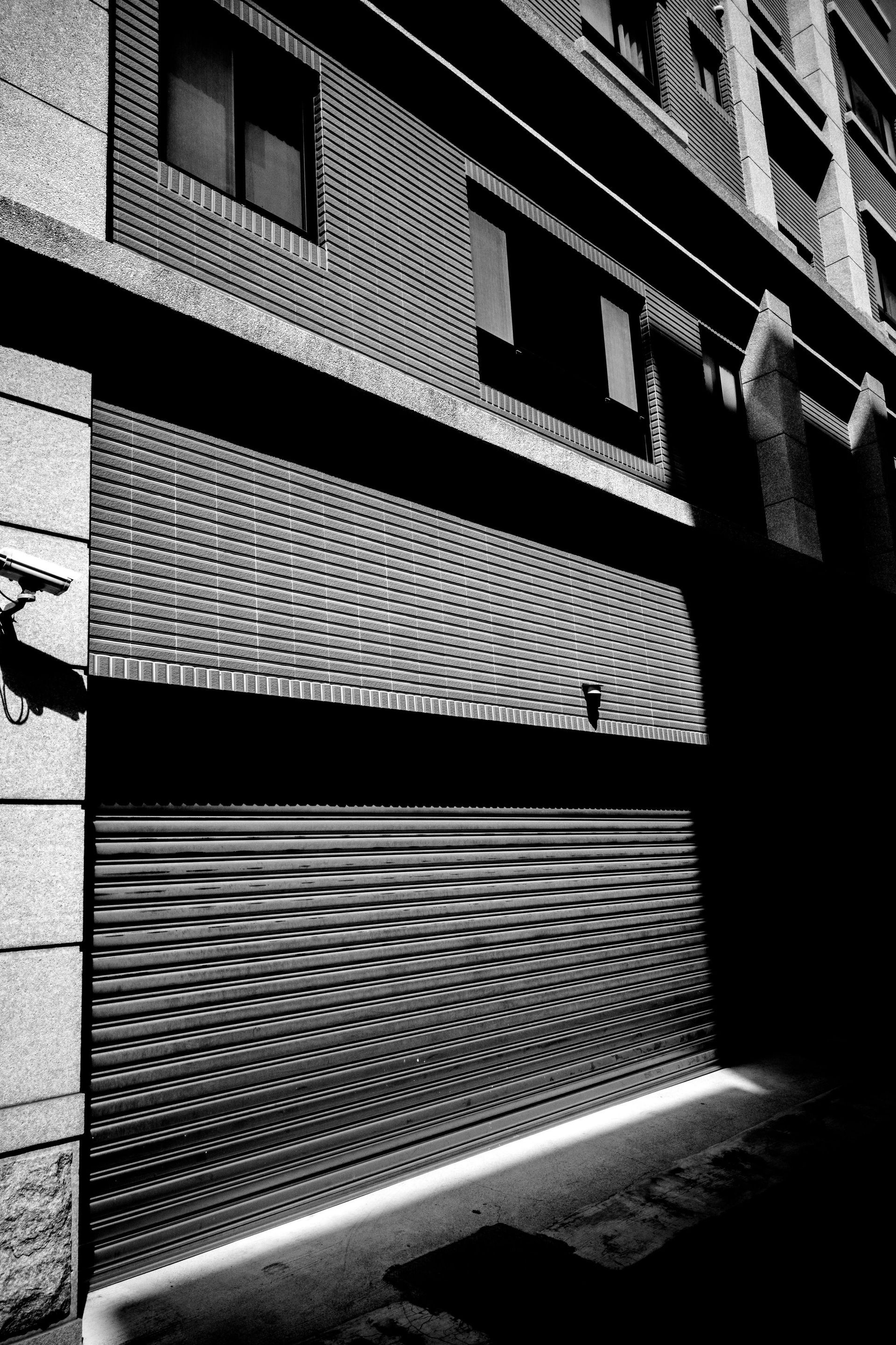 Black-and-white view of a narrow alley beside a brick building with shuttered windows and bright sunlight on the wall