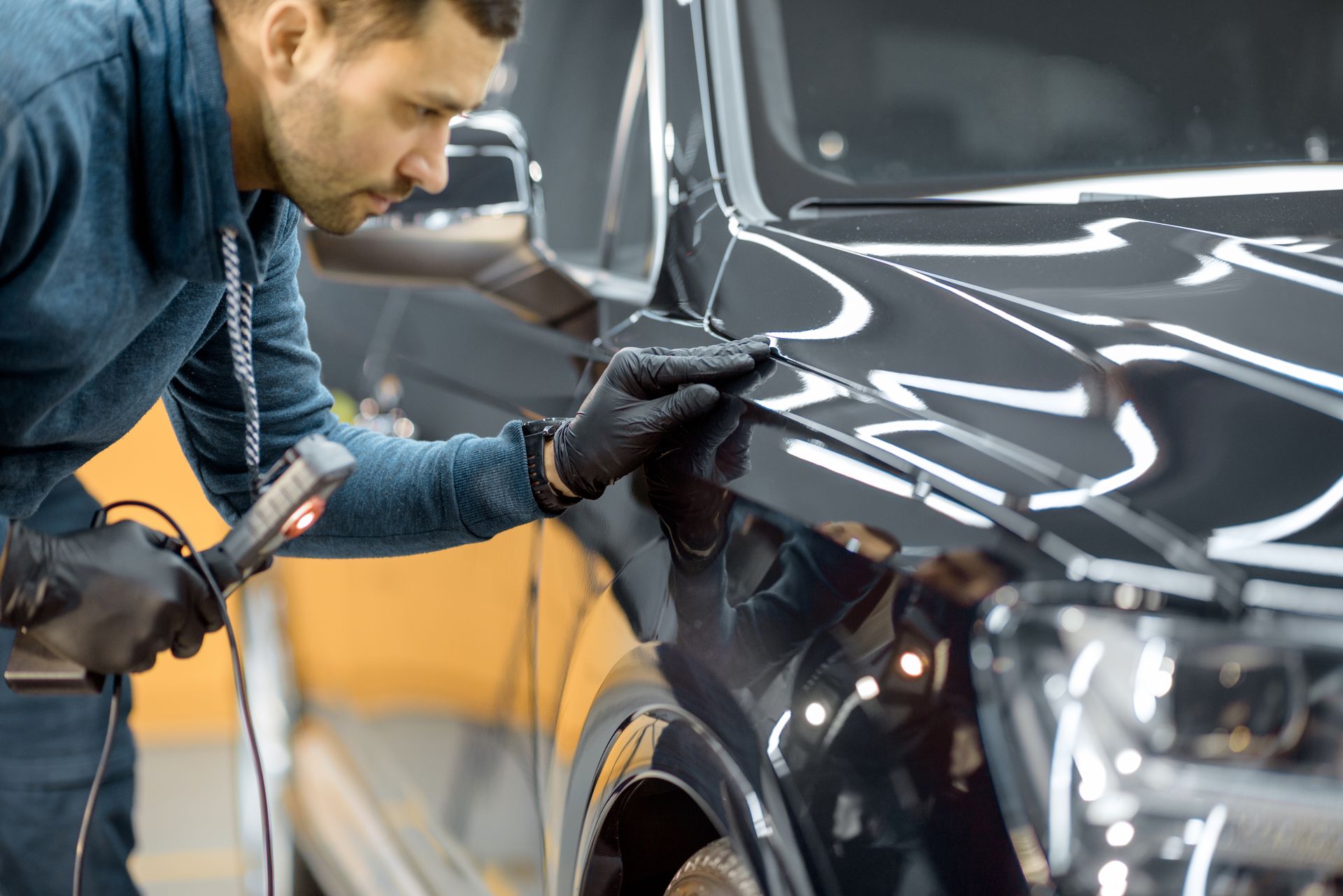 A worker in black gloves uses a handheld light to inspect the glossy black paint of a car in a workshop.
