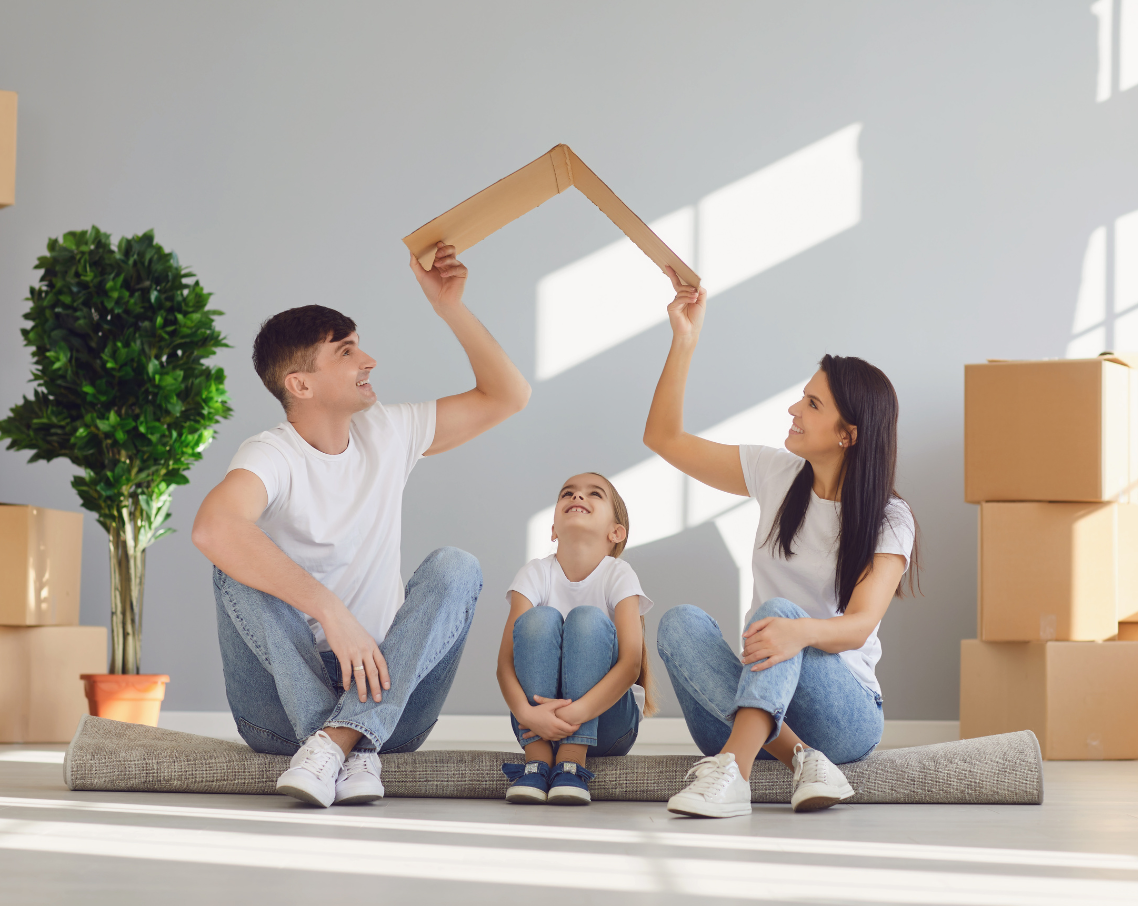 A family is sitting on the floor in a room holding a cardboard house.