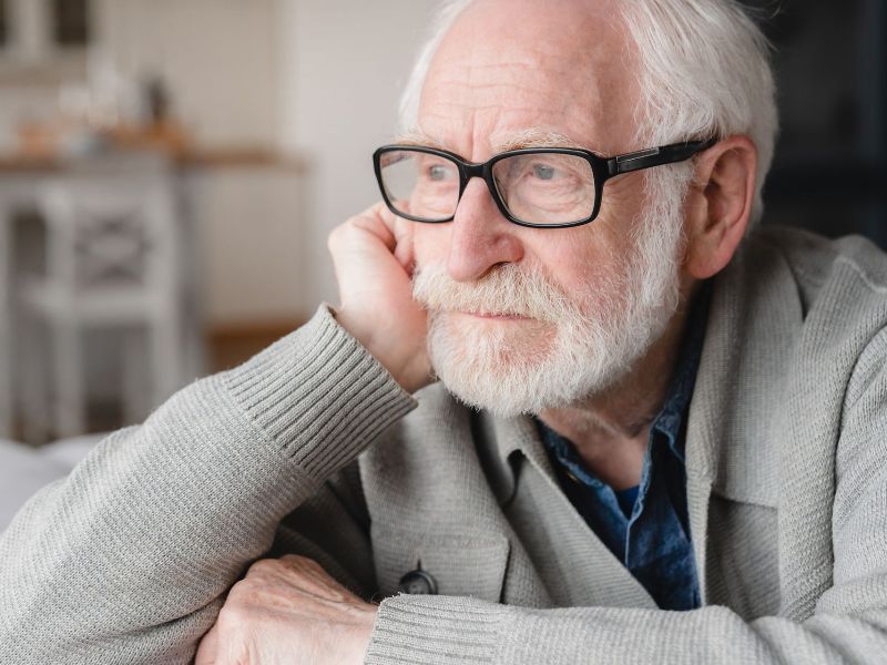 An elderly man with glasses and a beard is sitting on a couch.