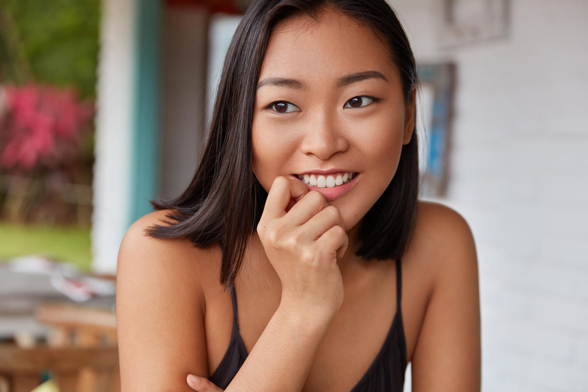 Woman with dark hair smiles, biting finger, wearing black tank top. Outdoors, blurred background.