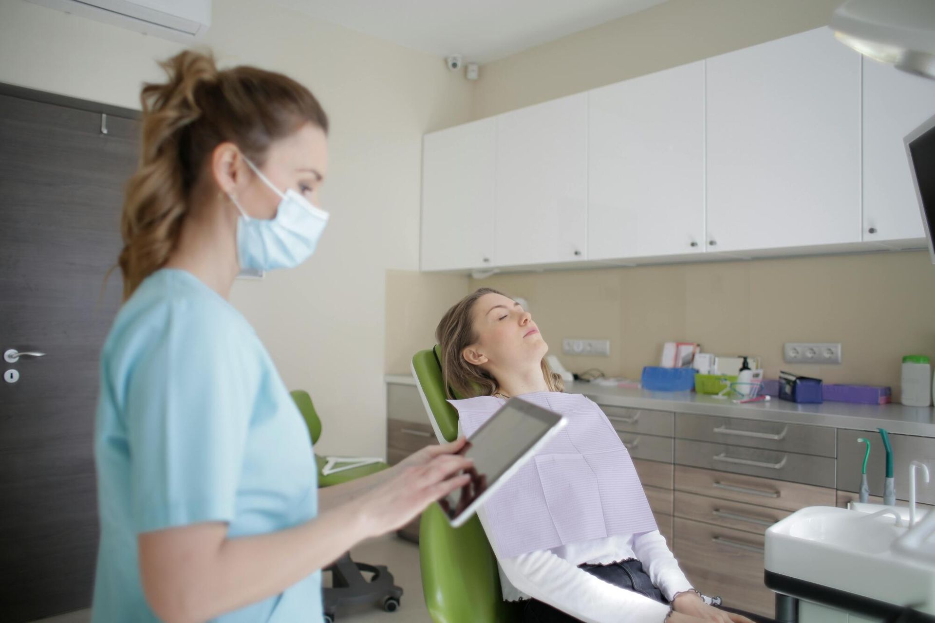 Dentist wearing a mask takes notes as a patient reclines in a dental chair in a bright clinic