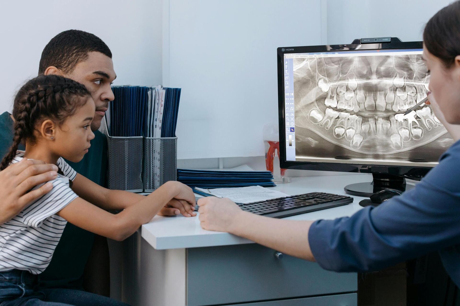 A dentist points to a dental x-ray on a monitor for a father and daughter in a dental office.