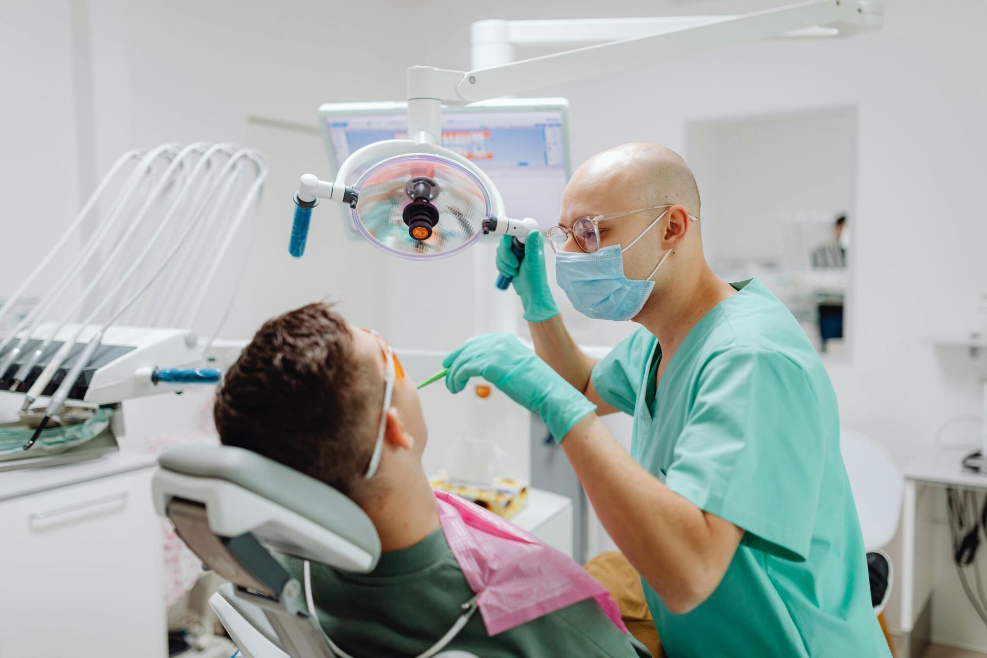 A dentist in scrubs, mask, and gloves examines a patient reclining in a dental chair under a professional exam light.