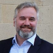 A man with a beard and a suit is smiling in front of a brick wall.