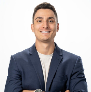 Man smiling, wearing a blue blazer and white t-shirt, arms crossed, studio backdrop.
