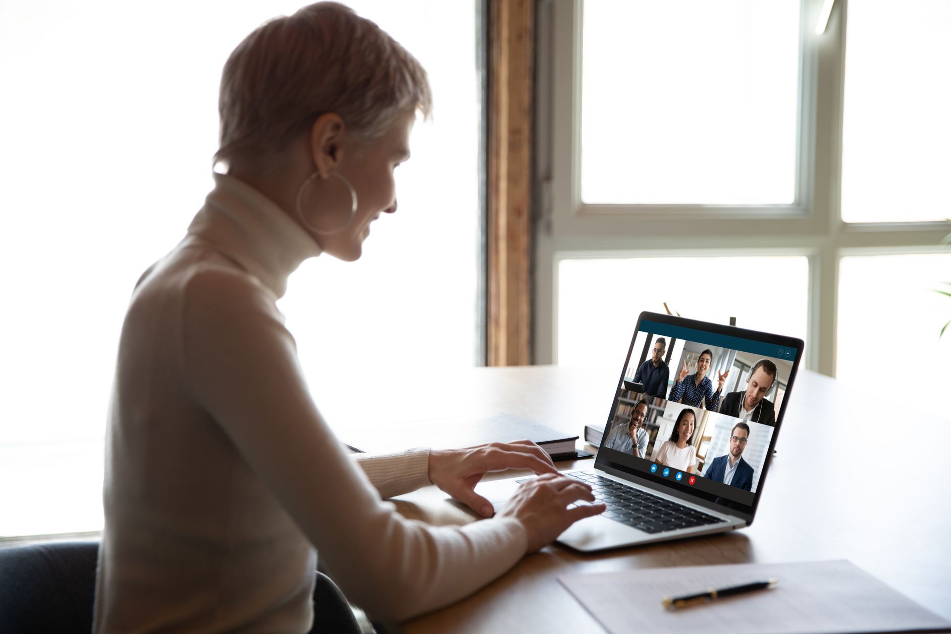 Woman in beige turtleneck types on laptop, attending video conference with colleagues. Bright room, neutral tones.