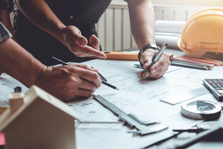 two employees discussing blue prints that are layed out over a table while a yellow hard hard sits to the side.